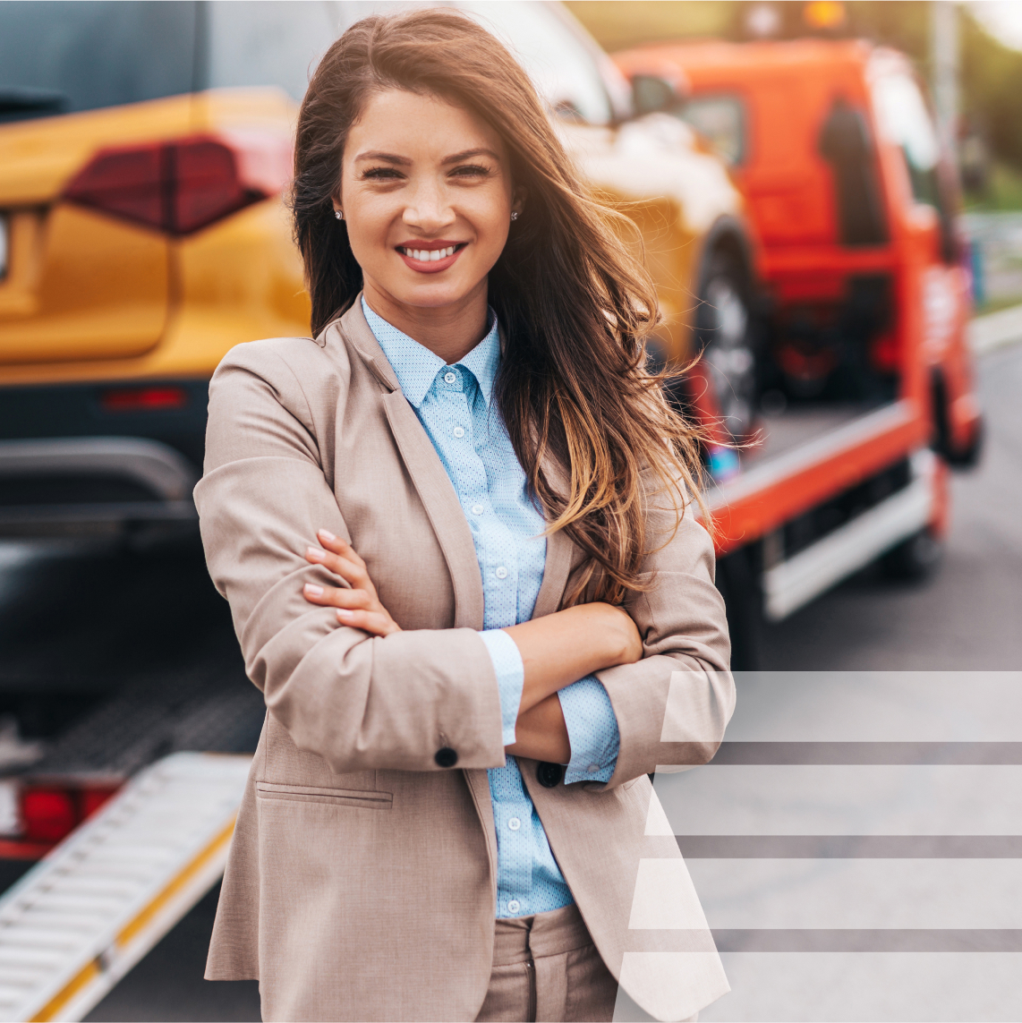 a woman smiling at the camera with crossed arms standing by a street. behind her a tow truck is hauling a car